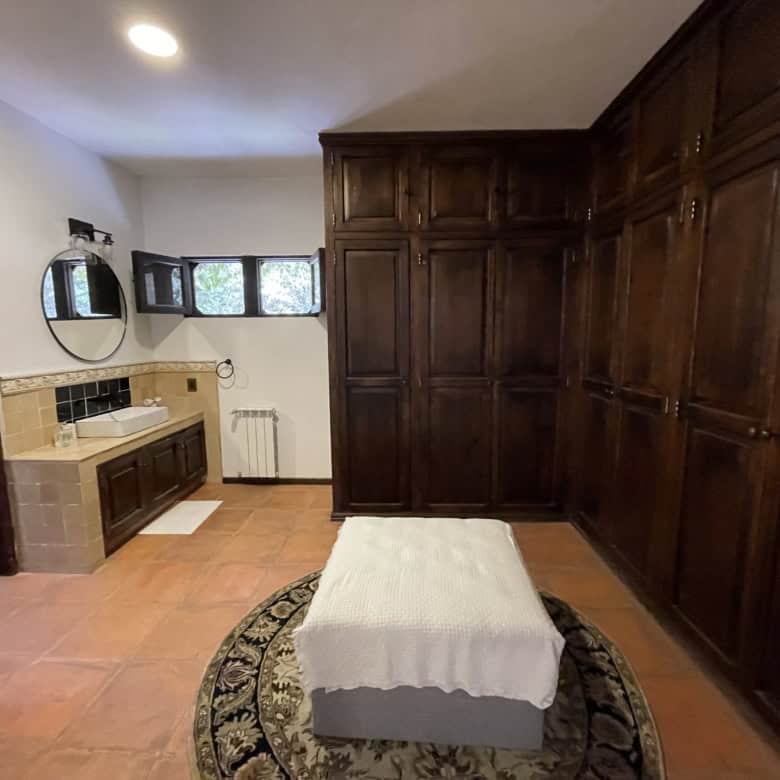 Spacious bedroom featuring large wooden wardrobes and a small bed, with a sink area and natural ligh.