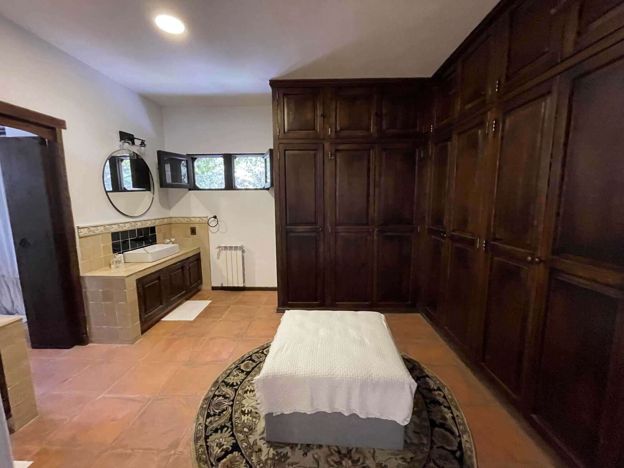 Spacious bedroom featuring large wooden wardrobes and a small bed, with a sink area and natural ligh.