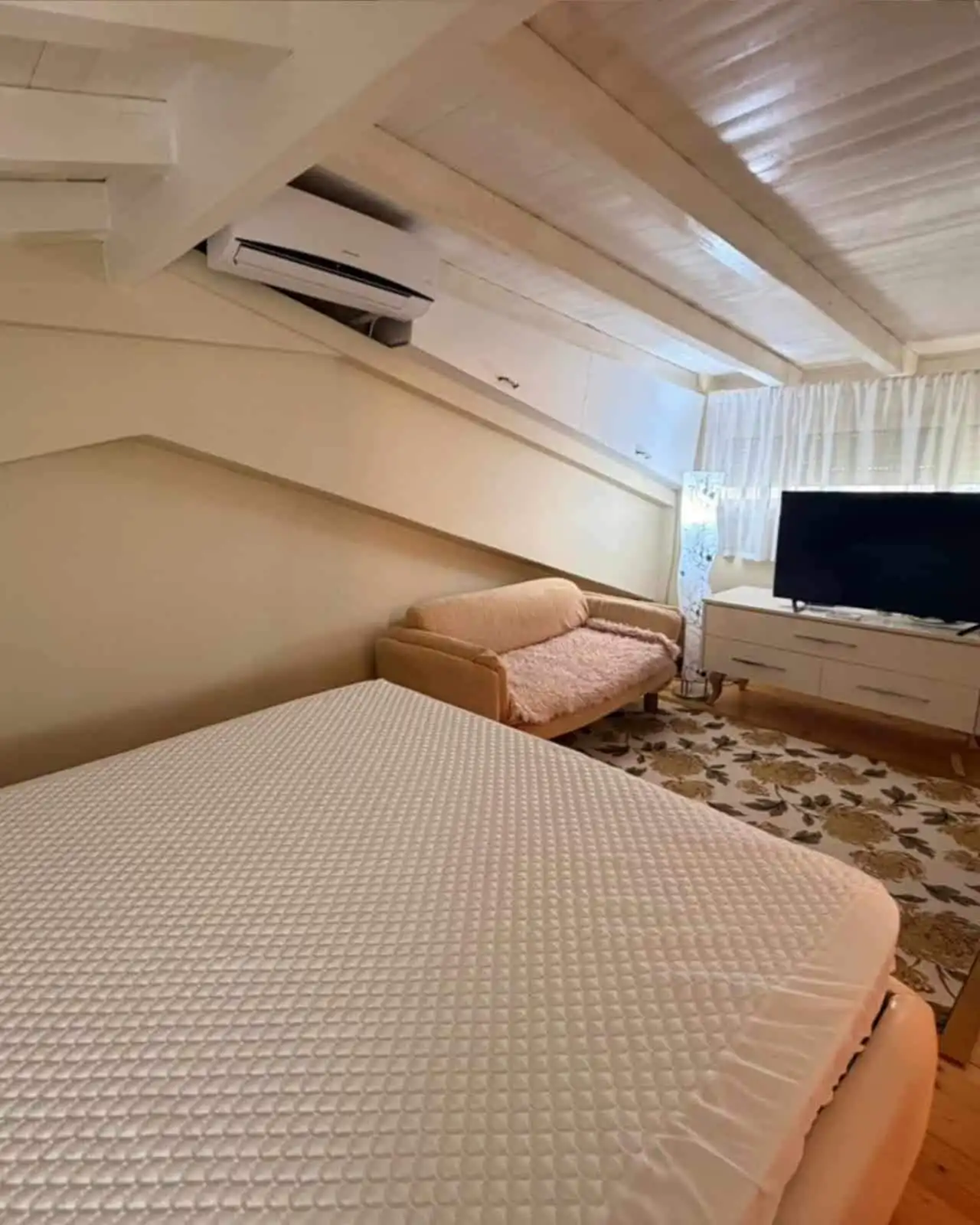 Attic bedroom featuring a bed, sofa, and TV in a bright, airy space with wooden ceiling and large wi.