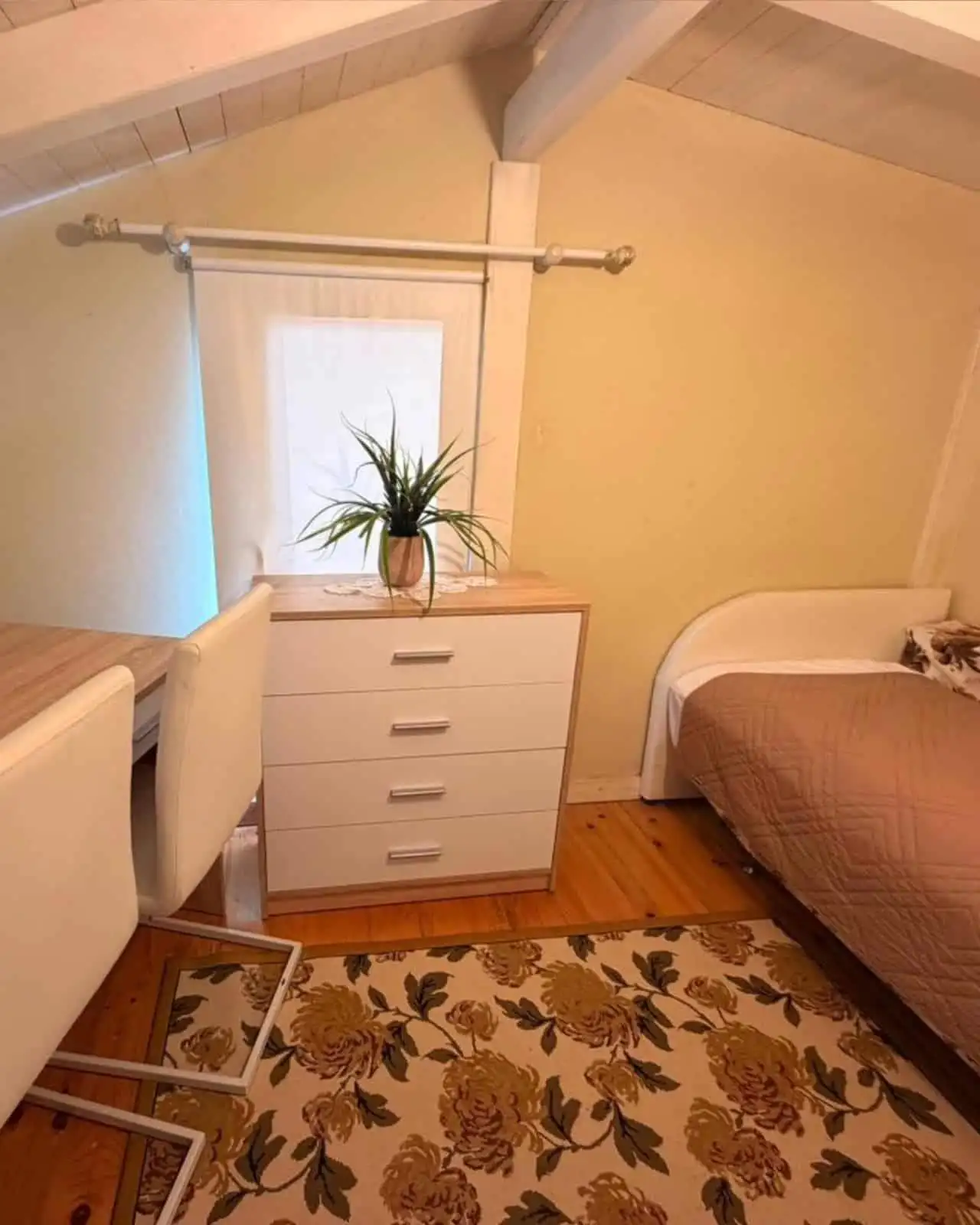 Bright bedroom featuring a sloped ceiling, a window with sheer curtains, and a simple dresser with a.