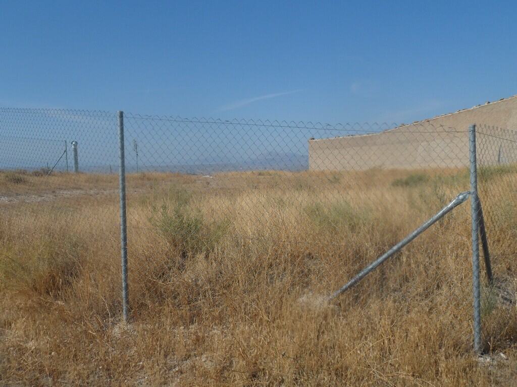 Fenced land near a cave house in Cuevas del Campo, Granada, with dry grass and a clear blue sky.