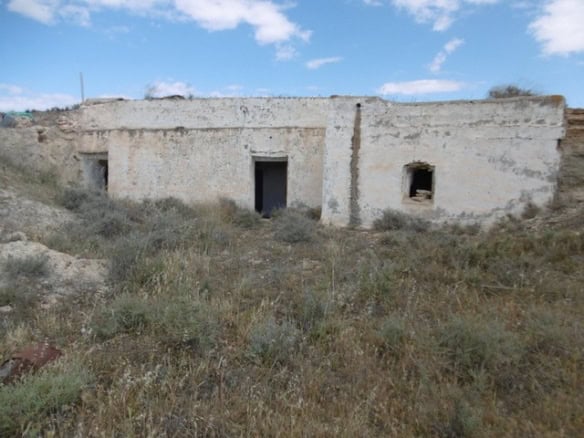 Cave house ruins in Benamaurel, Granada, with a clear sky and overgrown surroundings.