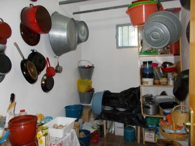 Rural kitchen with hanging pots and pans, shelves with kitchenware, and a small window providing nat.
