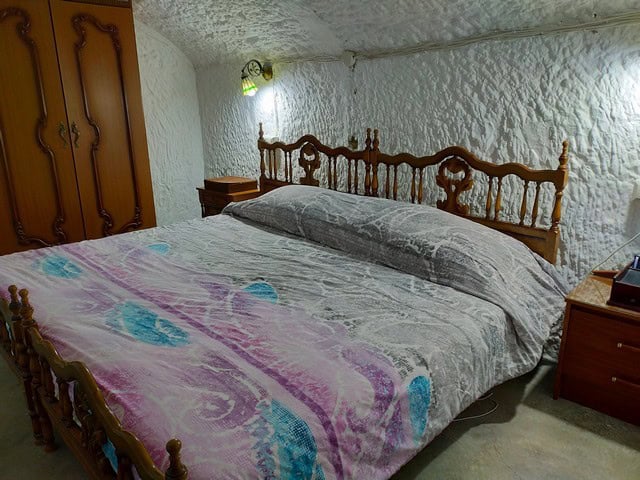 Cave house bedroom with rustic wooden bed and textured white walls in Benamaurel, Granada.