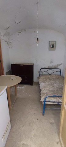Interior of a rustic cave house with a simple bed, small dresser, and minimal decor in La Alqueria,.