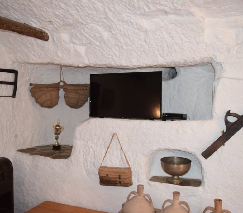 Cave house interior with traditional decor in Carramaiza, Granada, featuring a TV, rustic shelves, a.