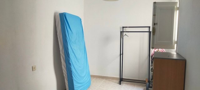 Interior view of a simple, unfurnished room with a mattress, clothing rack, and small cabinet.
