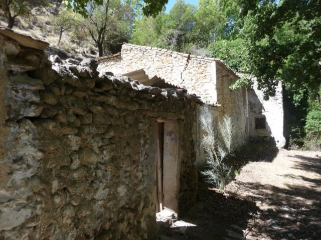 Rustic farm building with stone walls in Baza, Granada, surrounded by lush greenery and trees.