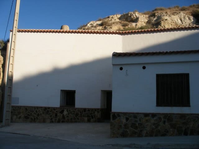 Traditional white cave house in Cuevas del Campo, Granada, with stone foundation and mountain backdr.