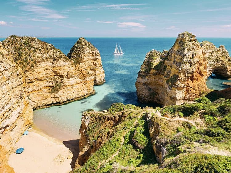 Cliffs and beach at Vale do Milho, Carvoeiro, Portugal, with a sailboat in the distance.
