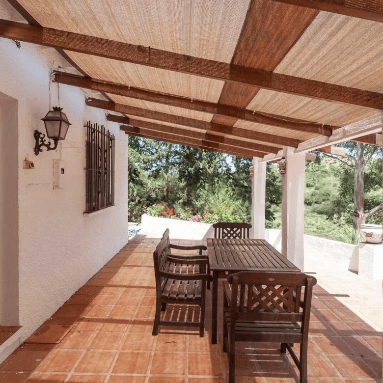Outdoor patio with wooden dining table and chairs under a shaded pergola. Surrounded by lush greener.