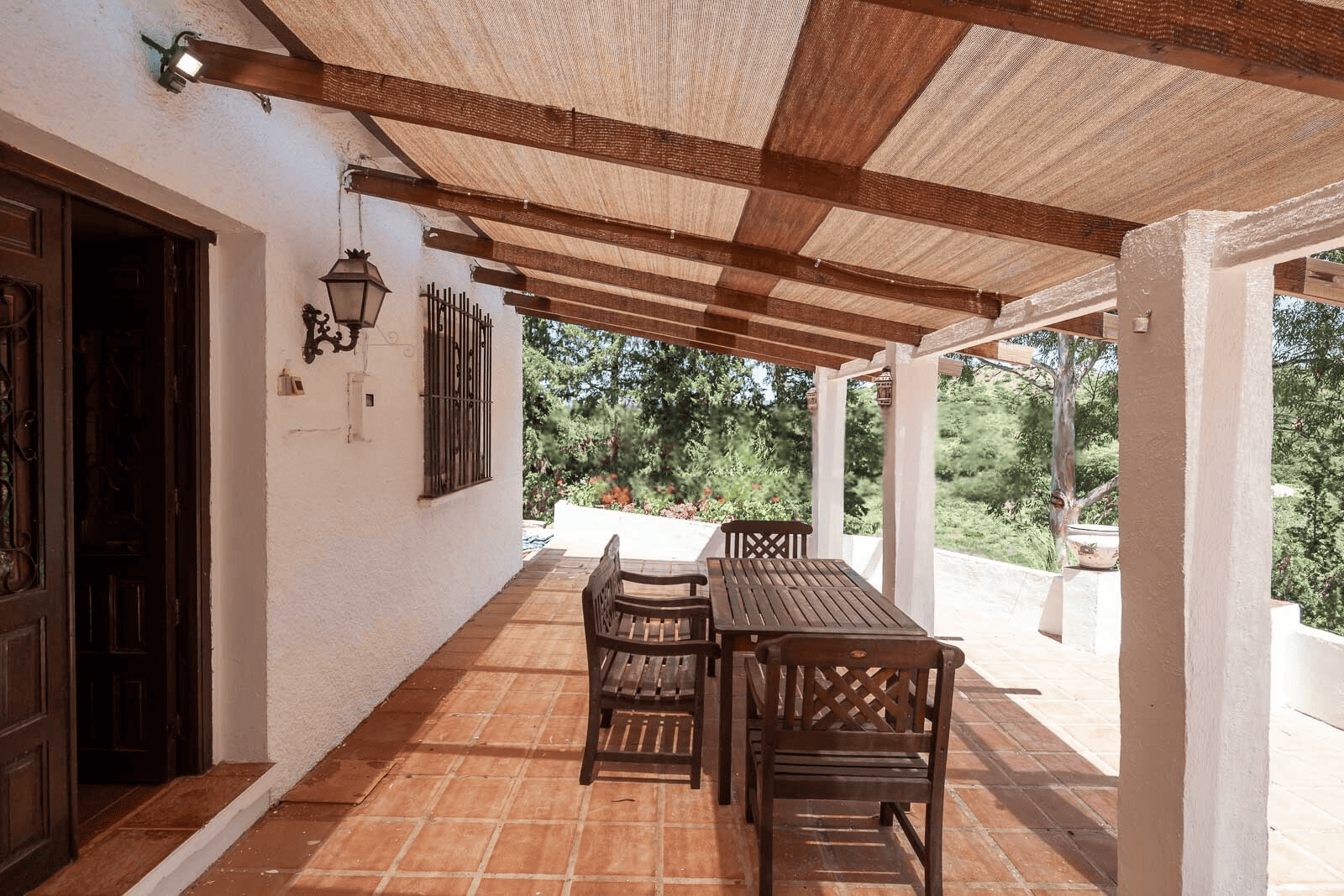 Outdoor patio with wooden dining table and chairs under a shaded pergola. Surrounded by lush greener.