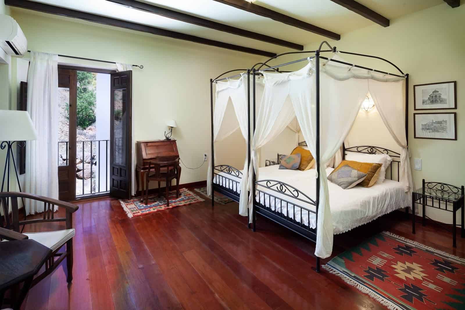 Cozy bedroom featuring a stylish canopy bed, wooden floors, and natural light from the balcony. Perf.