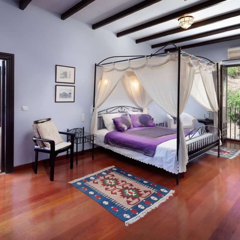 Luxury bedroom with a canopy bed, purple bedding, and balcony overlooking greenery.