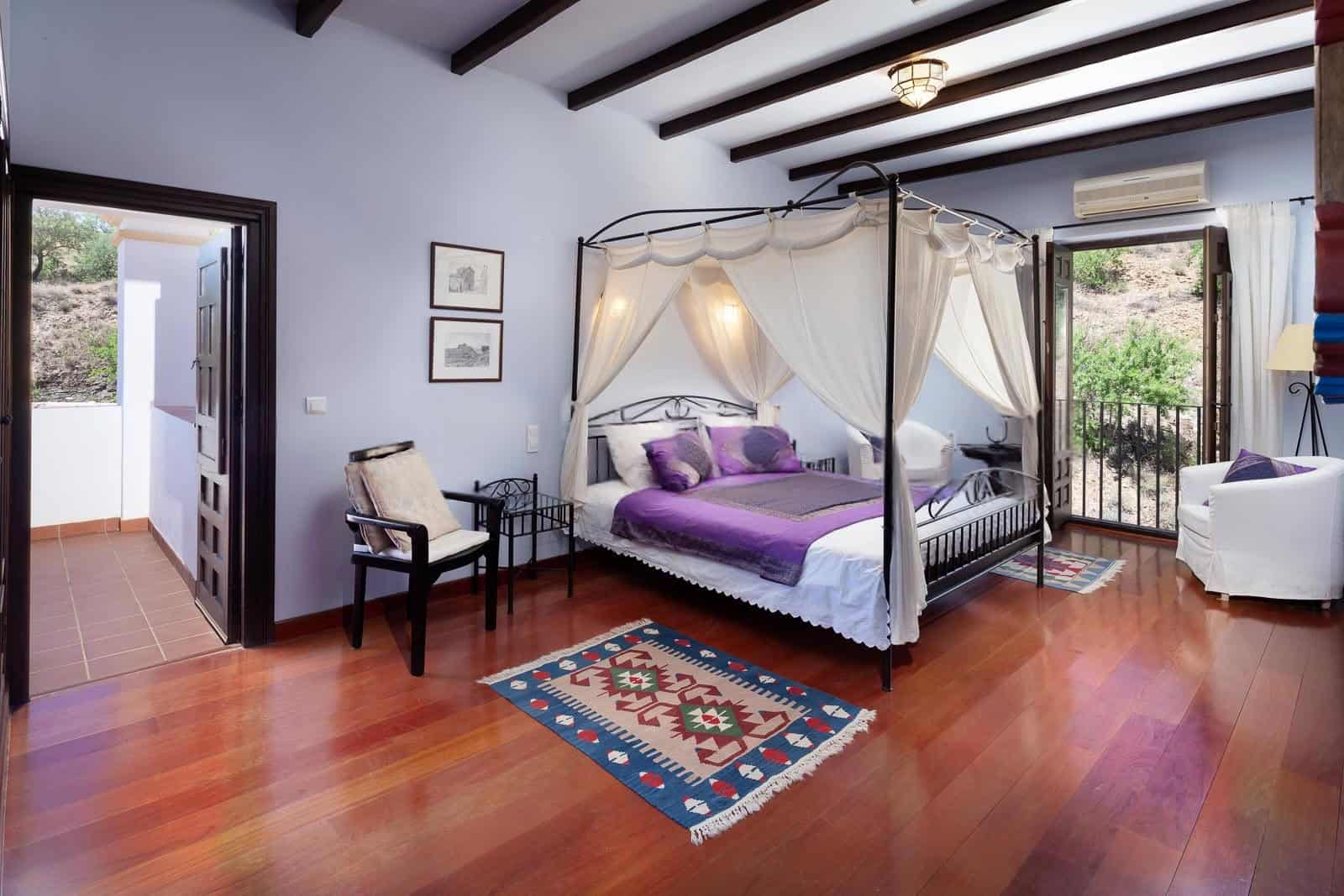 Luxury bedroom with a canopy bed, purple bedding, and balcony overlooking greenery.