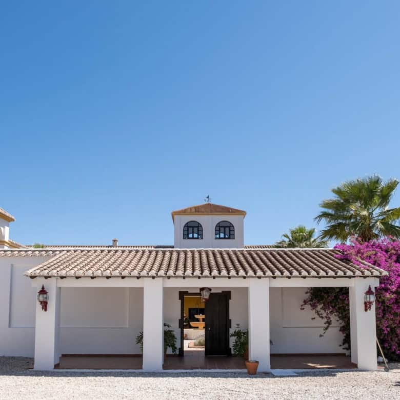 Luxury Spanish villa with garden, palm trees, and vibrant bougainvillaea under a clear blue sky.