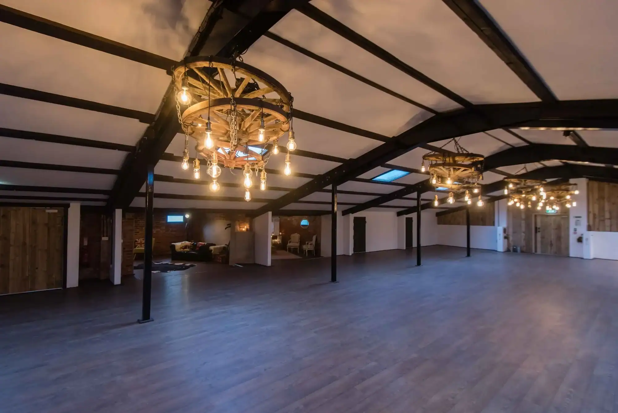 Spacious industrial-style event hall with wooden accents and unique chandeliers. Bright, open interior with rustic lighting fixtures and high ceilings.