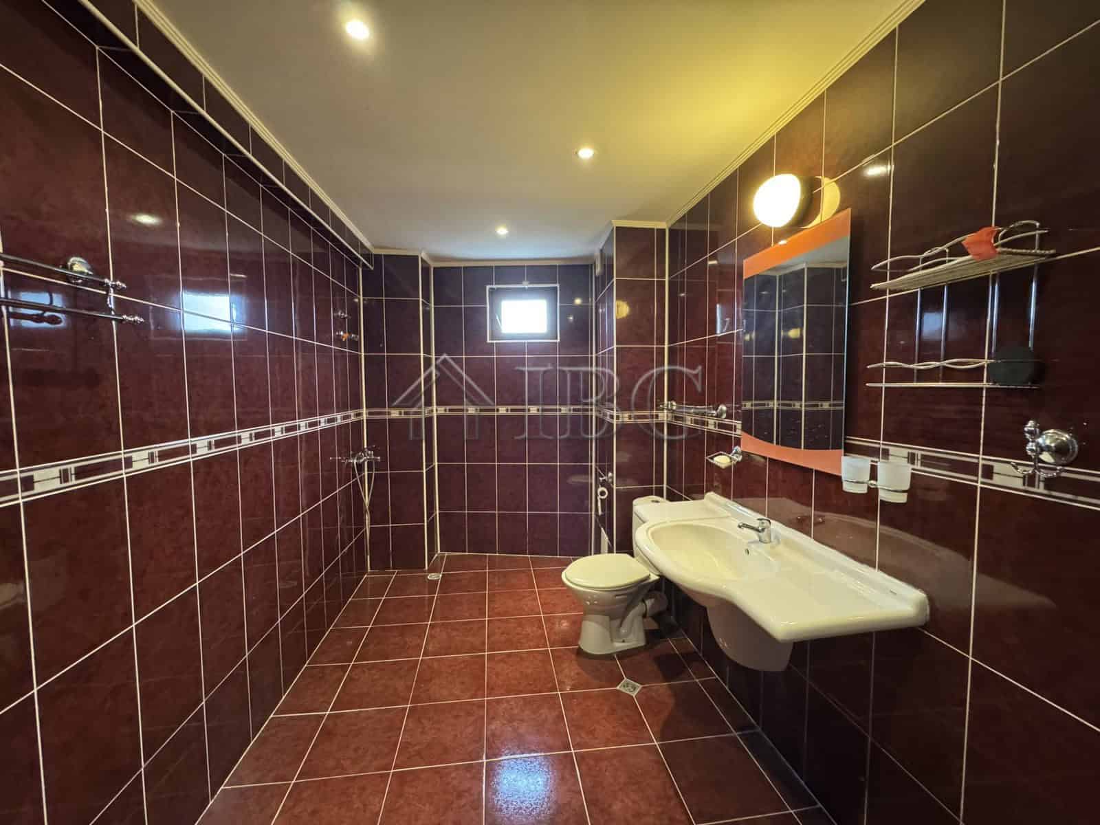 Modern bathroom featuring dark tiled walls and floor, with a white sink, toilet, and ample lighting.