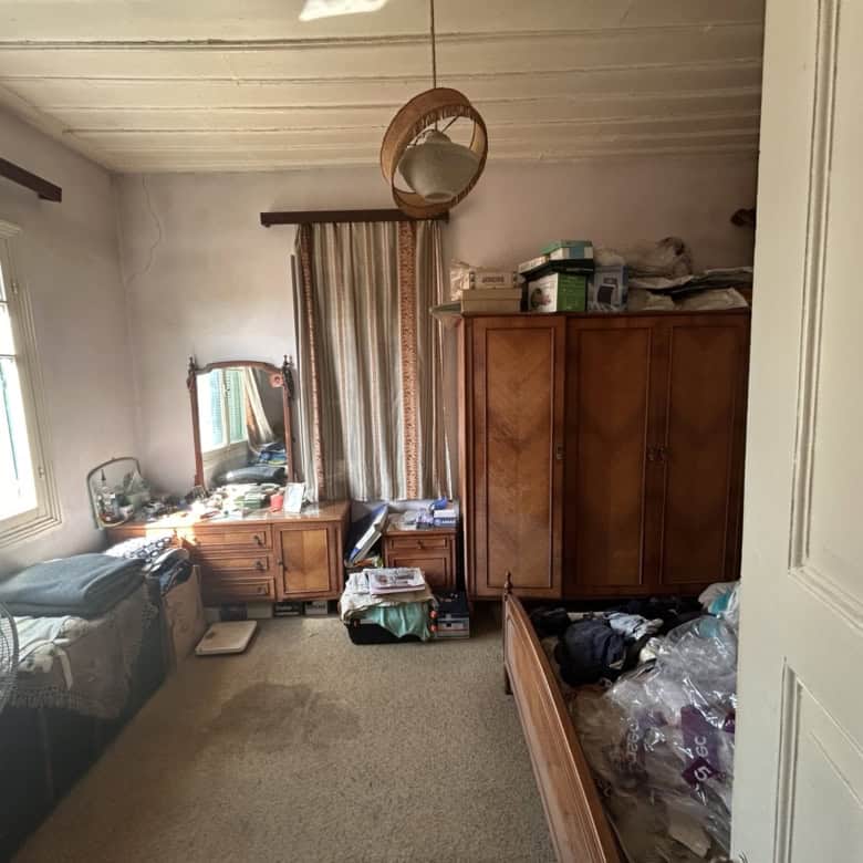 Cozy bedroom featuring wooden wardrobe, dresser, and a bed with cluttered items. Natural light from.