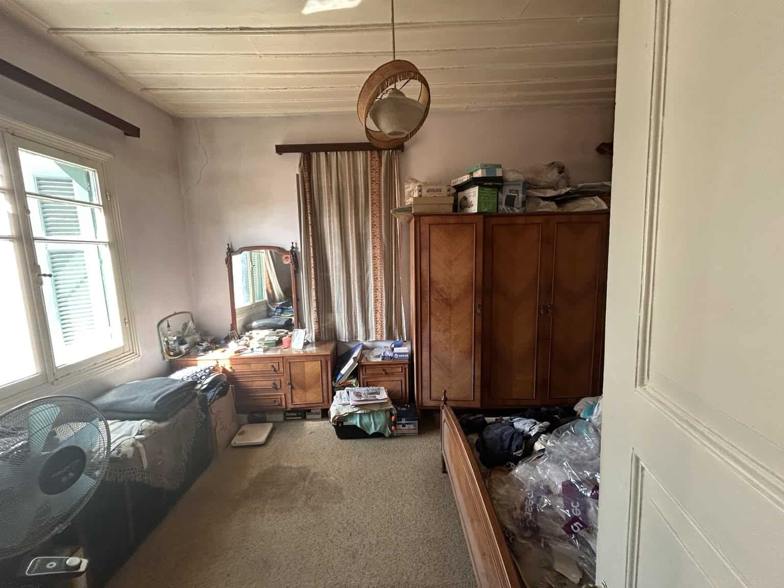 Cozy bedroom featuring wooden wardrobe, dresser, and a bed with cluttered items. Natural light from.