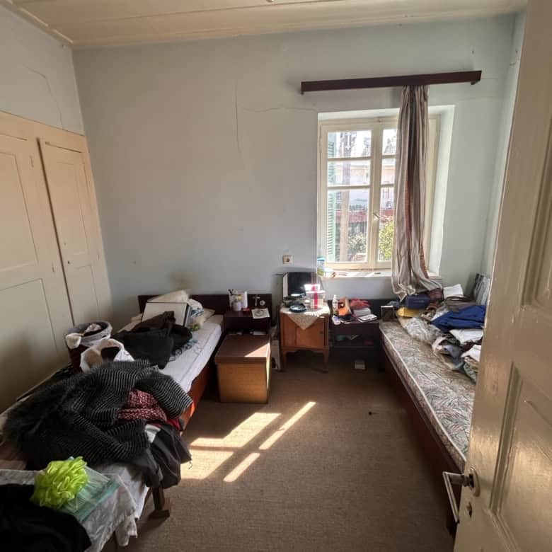 Bright bedroom with two single beds, cluttered with clothes and personal items, illuminated by sunli.