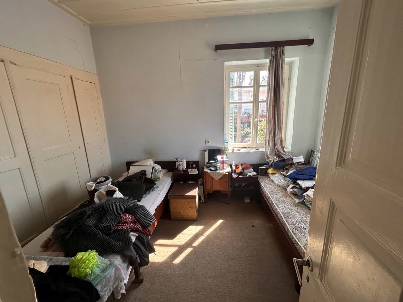 Bright bedroom with two single beds, cluttered with clothes and personal items, illuminated by sunli.
