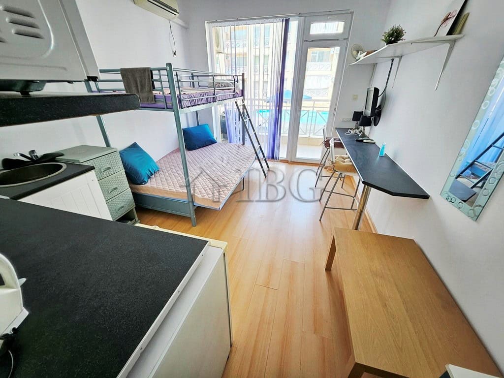Bright studio apartment with balcony overlooking pool and sunny beach scenery.
