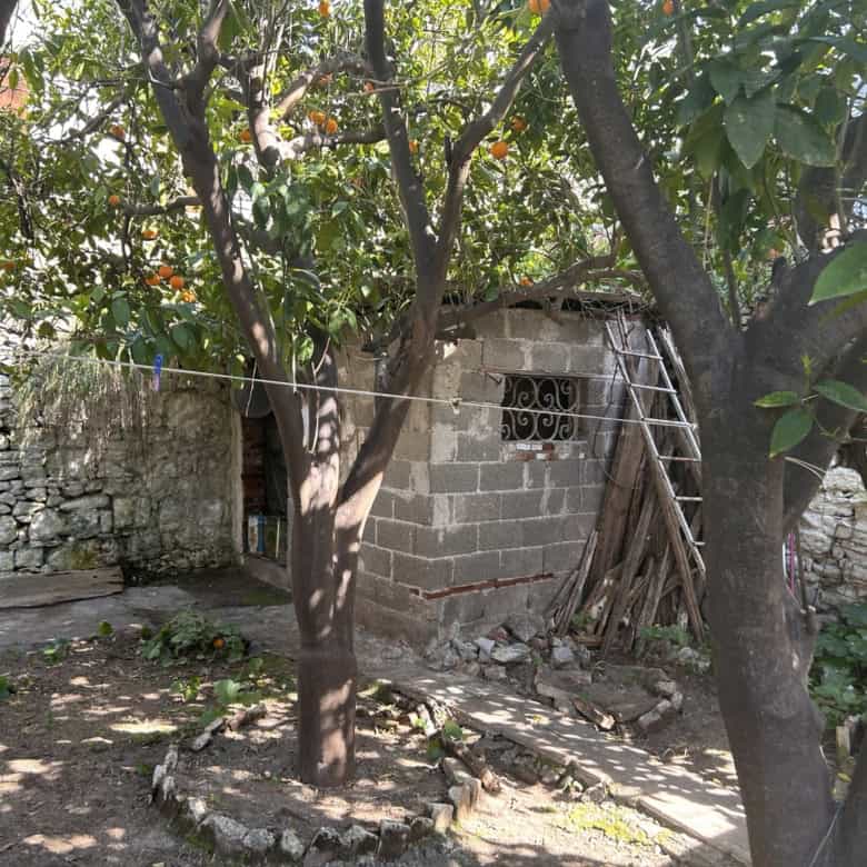 Small rustic house with trees and stone wall in a rural setting, showcasing traditional construction.