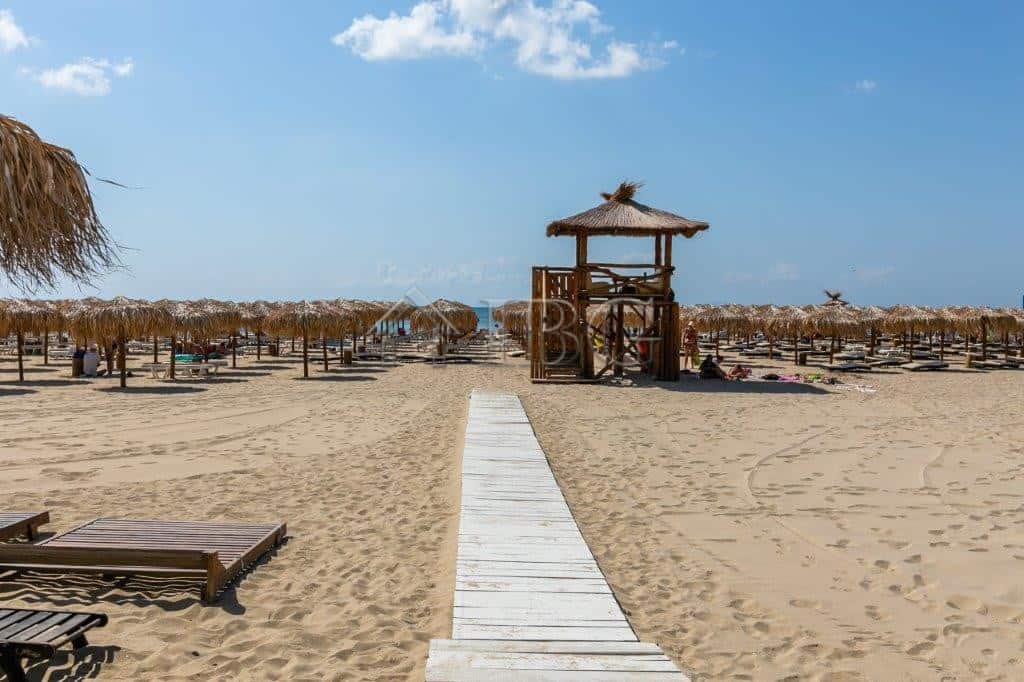 Beach scene with sun loungers, straw umbrellas, and a wooden walkway leading to the sea.