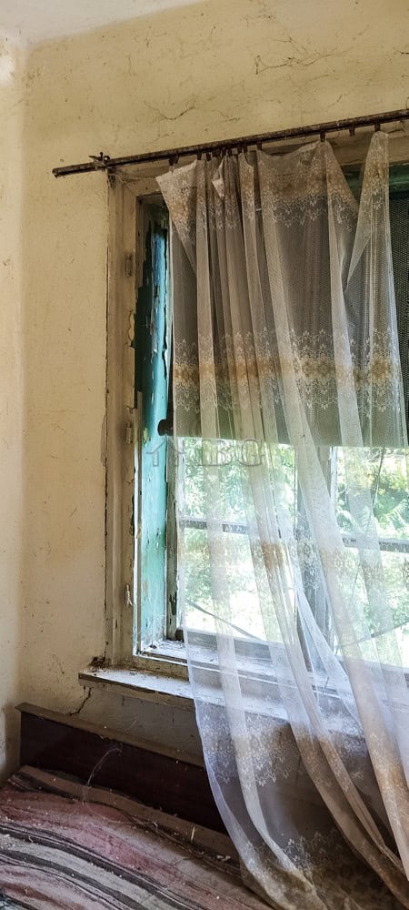 Old house interior with sheer curtains and a view outside through a worn window.