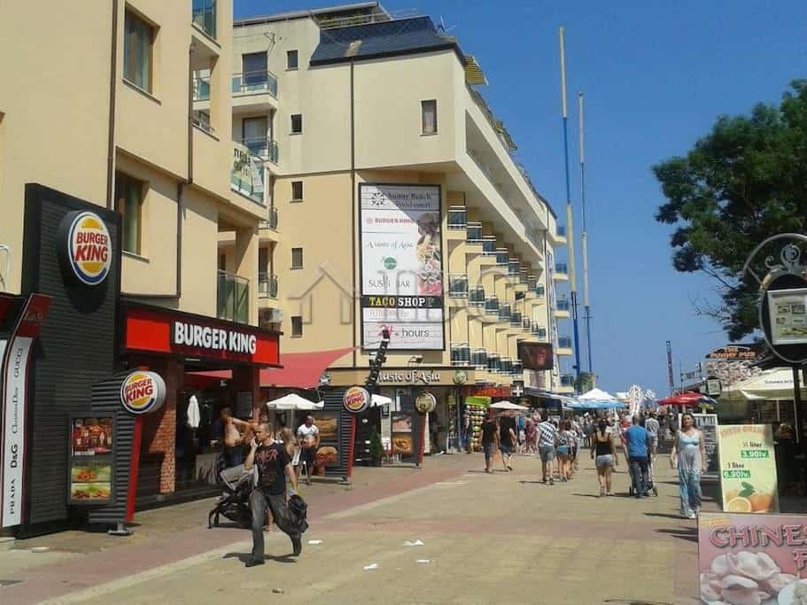 Bright street view of Kaya, Sunny Beach with a studio apartment building and commercial shops.