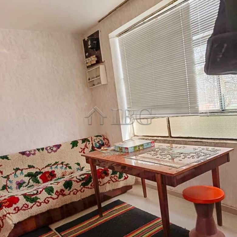 Bright living space featuring a large window, floral sofa cover, and a wooden table with colorful ru.