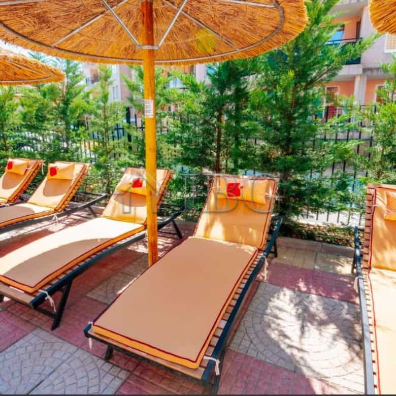 Sunbeds with umbrellas on a sunny terrace in Sunny Beach, surrounded by greenery and modern building.