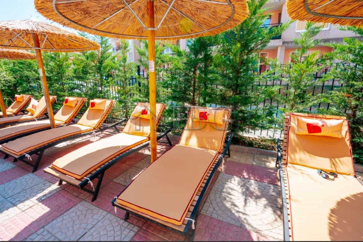 Sunbeds with umbrellas on a sunny terrace in Sunny Beach, surrounded by greenery and modern building.