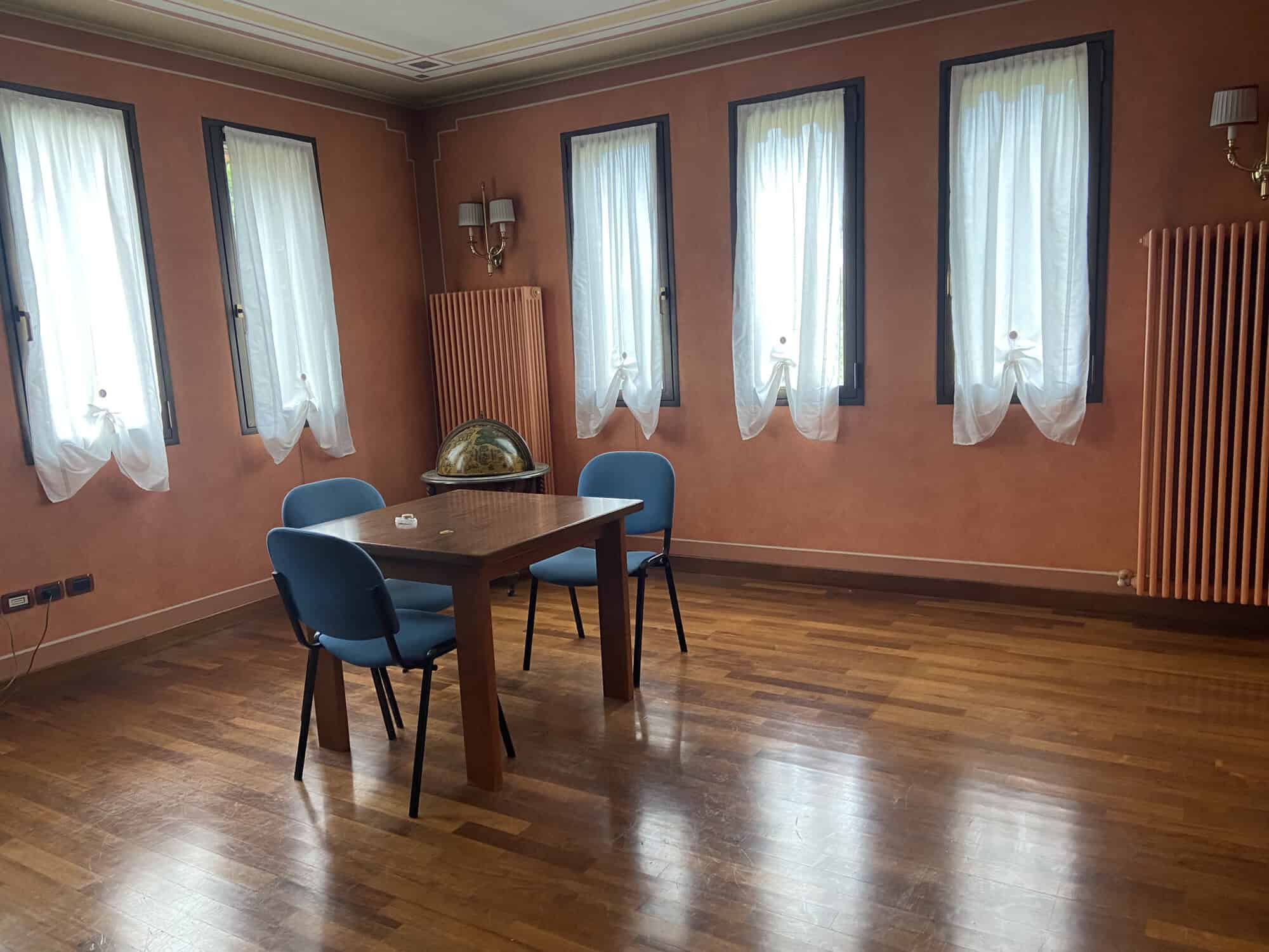 Spacious dining room with wooden floors, four windows with white curtains, and a wooden table with f.