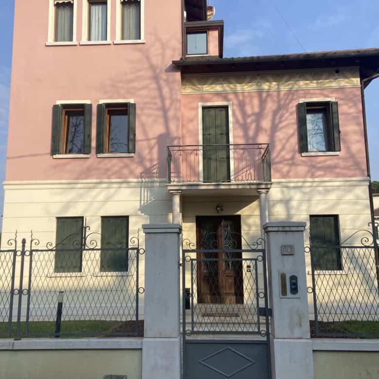 Elegant pink and beige villa with balcony, surrounded by a secure wrought iron gate and lush greener.