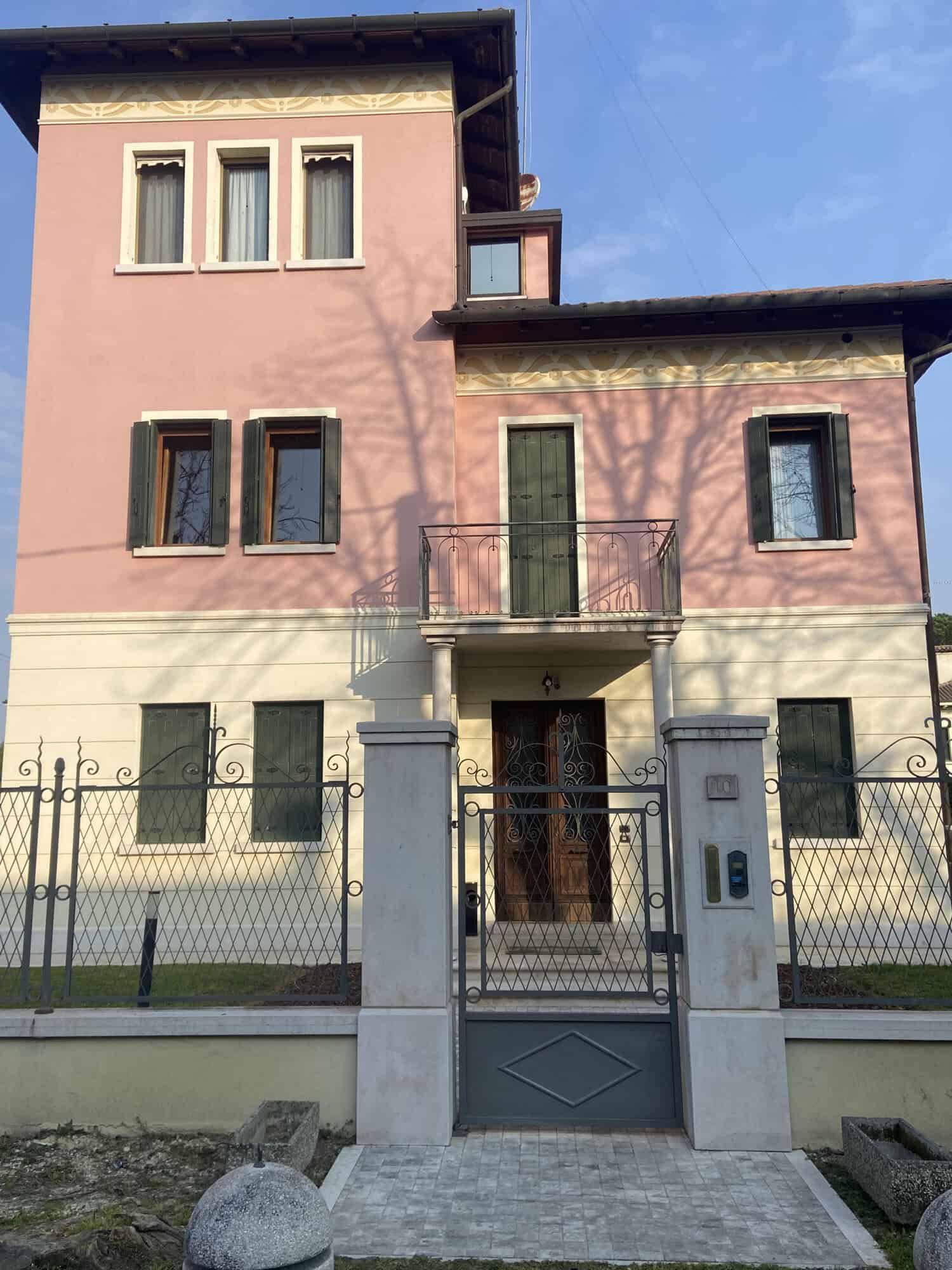 Elegant pink and beige villa with balcony, surrounded by a secure wrought iron gate and lush greener.