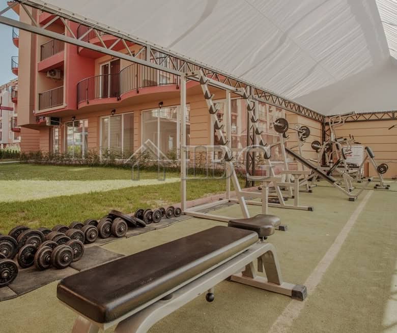 Outdoor gym area with fitness equipment and weights in a garden setting.
