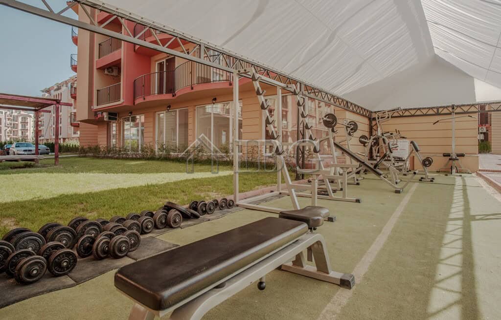 Outdoor gym area with fitness equipment and weights in a garden setting.