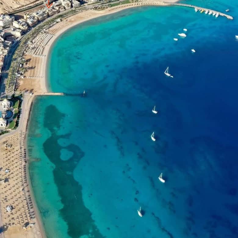 Beach and marina with sailboats in clear blue waters, sunny weather, and coastal cityscape.