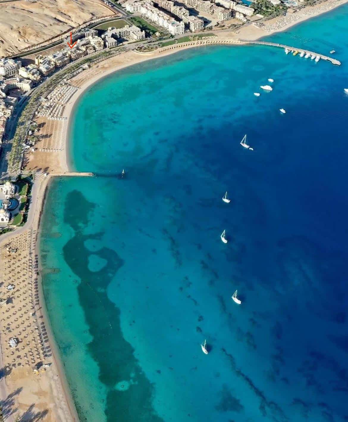 Beach and marina with sailboats in clear blue waters, sunny weather, and coastal cityscape.