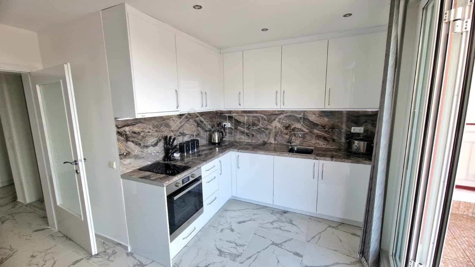 Modern white kitchen with marble countertops and large glass door opening to a balcony.