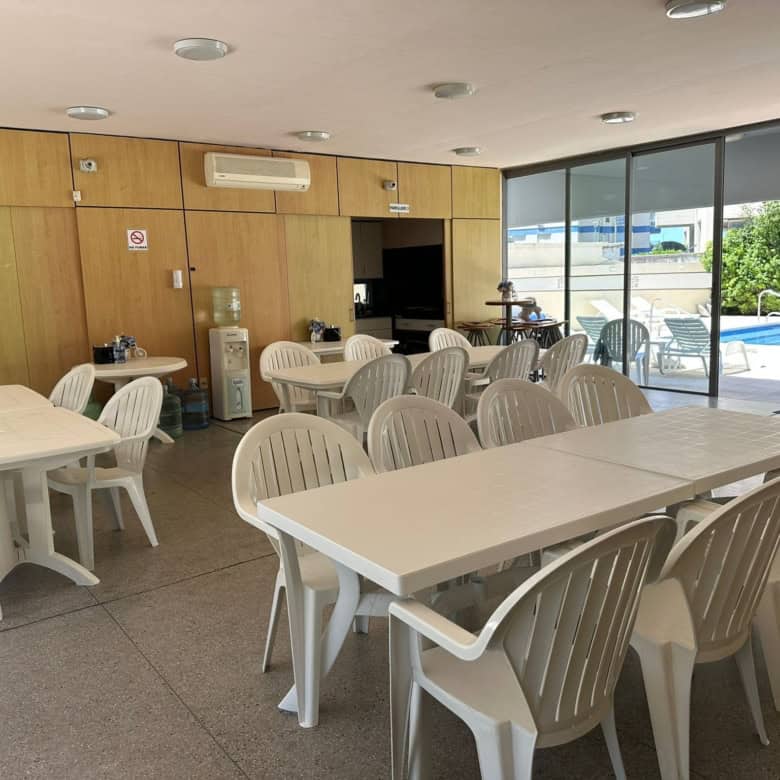 Indoor lounge area with white plastic chairs and tables, overlooking pool outside.