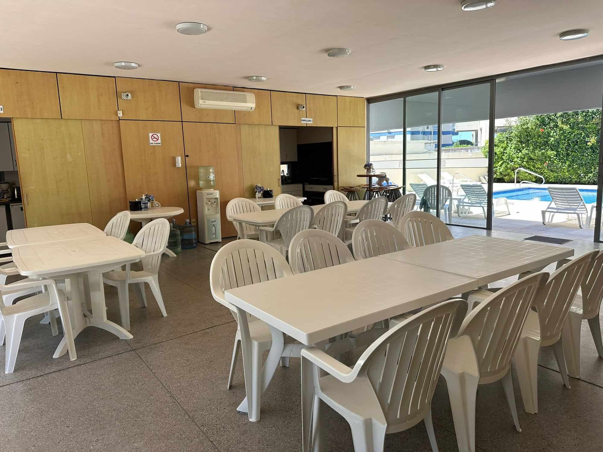 Indoor lounge area with white plastic chairs and tables, overlooking pool outside.