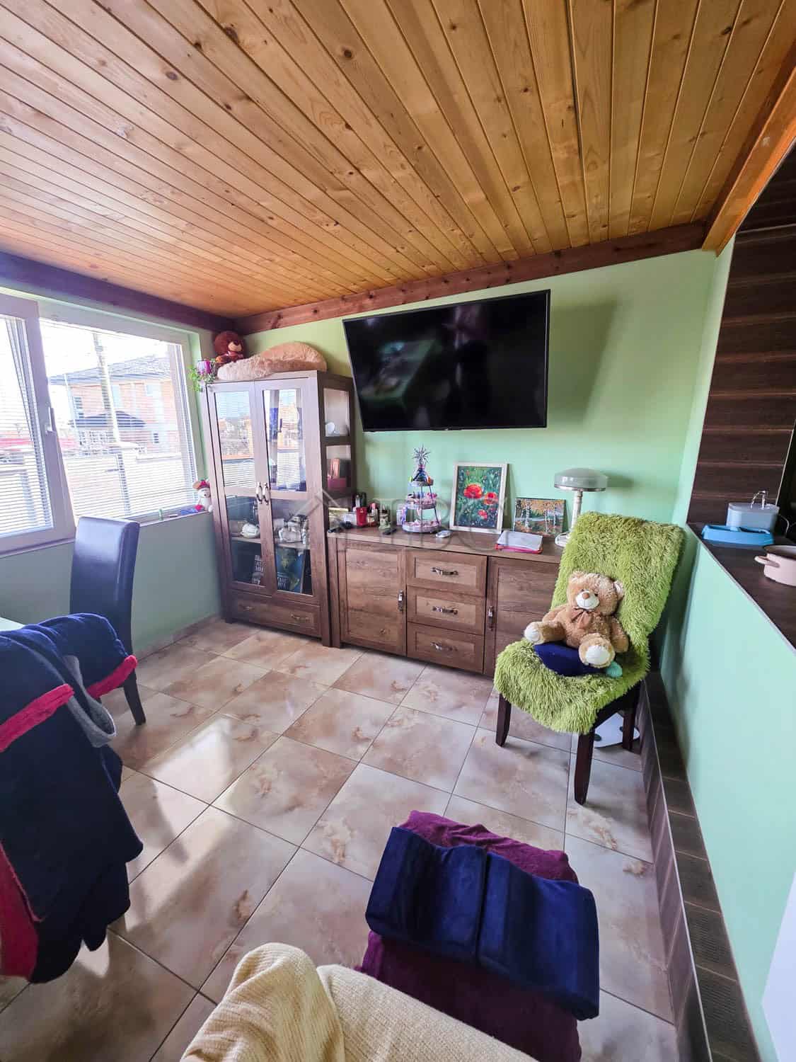 Bright living room with a wall-mounted TV, wooden cabinet, and cozy green armchair with teddy bear.
