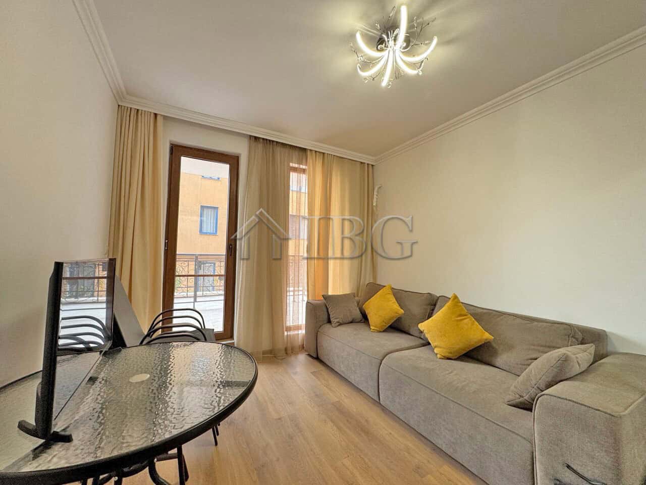 Spacious living room with sofa and yellow cushions, large windows with curtains, and a modern chande.