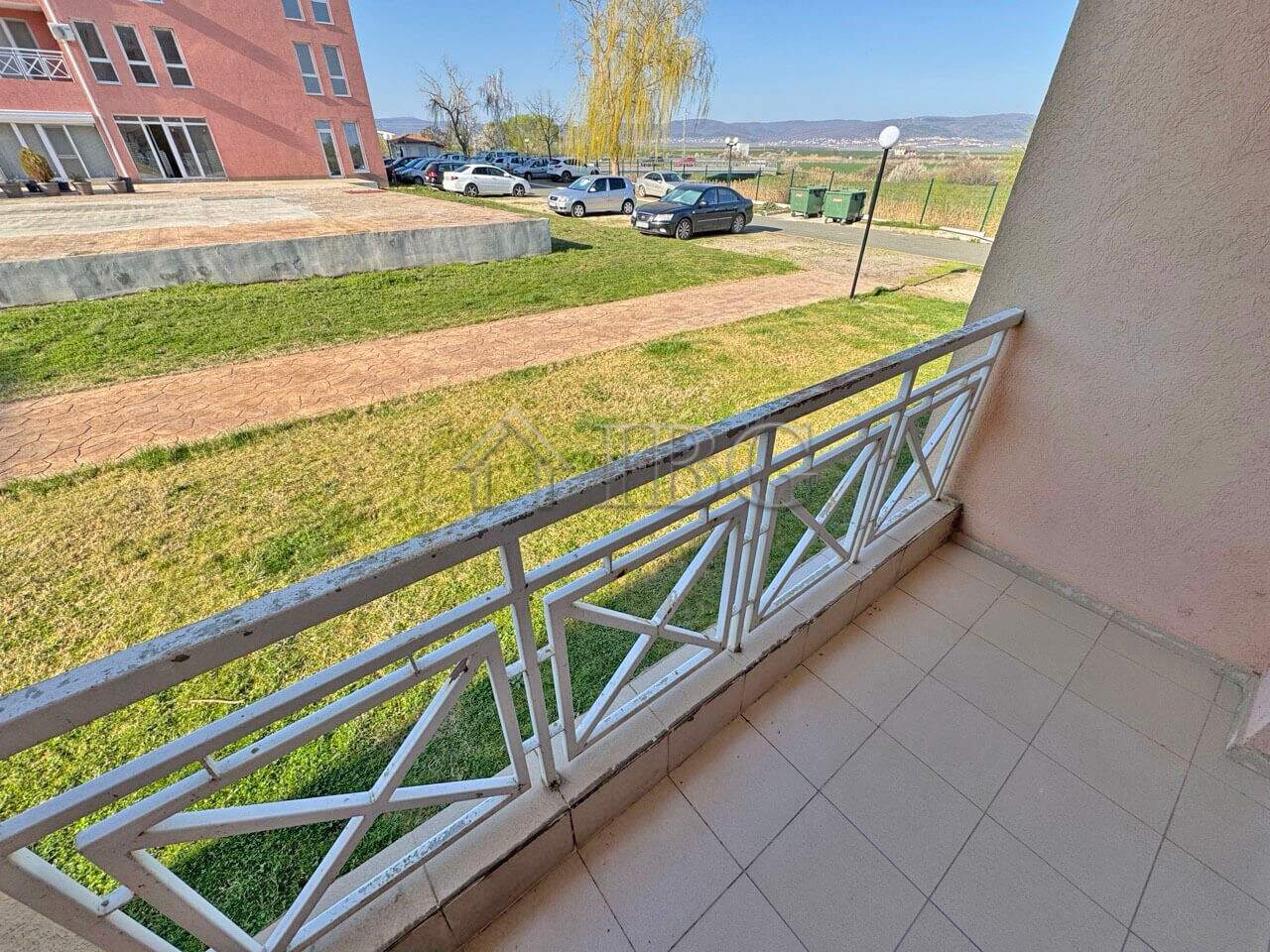 Ground-floor apartment with balcony overlooking green yard and parking area.